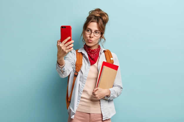 Hermosa mujer caucásica de cabello oscuro hace retrato selfie con teléfono celular, mantiene los labios doblados, lleva cuaderno y mochila en la espalda