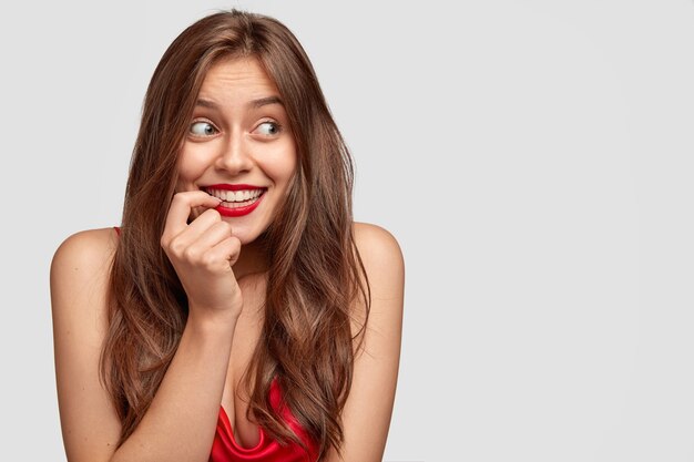 Hermosa mujer caucásica con cabello oscuro, sonrisa con dientes, usa maquillaje mínimo y lápiz labial rojo, mira con alegría a un lado, modelos contra la pared blanca con espacio libre para su texto promocional