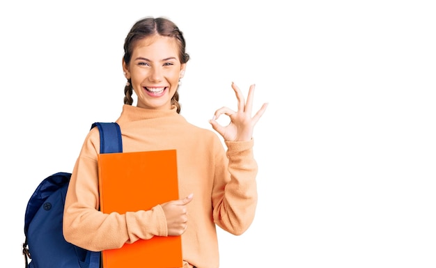 Hermosa mujer caucásica con cabello rubio con mochila de estudiante y sosteniendo un libro haciendo el signo de ok con los dedos sonriendo amigable gesticulando excelente símbolo