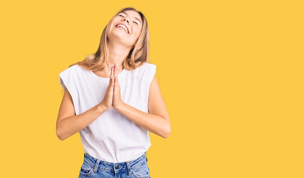 Foto gratuita hermosa mujer caucásica con cabello rubio usando camiseta blanca casual rogando y rezando con las manos juntas con expresión de esperanza en la cara muy emocional y preocupada. mendicidad.