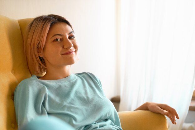 Hermosa mujer caucásica joven encantadora con peinado rosado bob y piercing facial relajante en casa, sentado cómodamente en el sofá, mirando a la cámara con una linda sonrisa