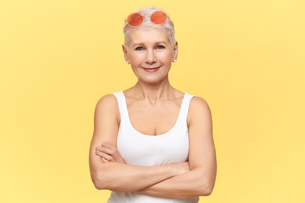 Hermosa mujer caucásica jubilada con gafas de sol rosas redondas y camiseta blanca, cruzando los brazos sobre el pecho, con expresión facial segura.