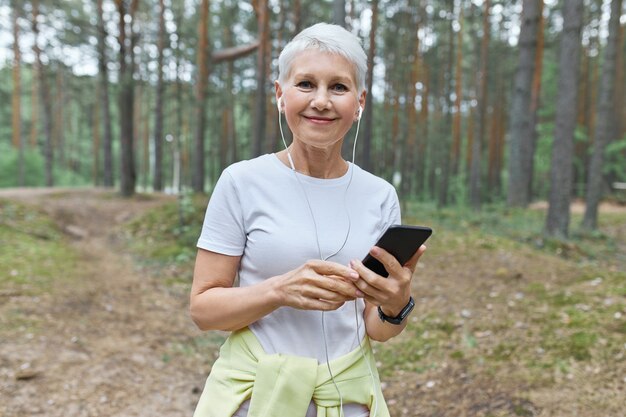 Hermosa mujer caucásica de mediana edad con cabello corto disfrutando de la mañana de verano al aire libre, va a hacer ejercicio cardiovascular, eligiendo pistas de música en el teléfono móvil