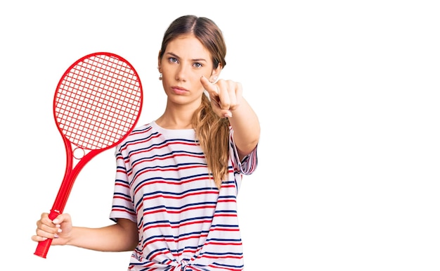 Hermosa mujer caucásica con el pelo rubio jugando al tenis sosteniendo la raqueta apuntando con el dedo a la cámara y a ti, gesto de confianza que parece serio