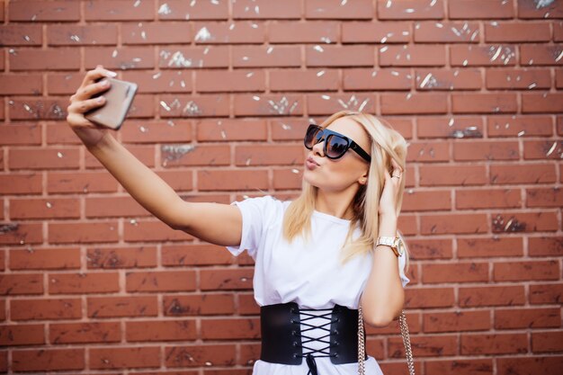 Hermosa mujer caucásica rubia de pie delante de la pared de ladrillo y hacer selfie.