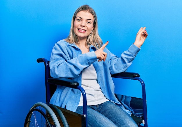 Hermosa mujer caucásica sentada en silla de ruedas sonriendo y mirando a la cámara apuntando con dos manos y dedos a un lado