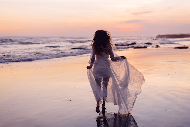 Hermosa mujer caucásica sola en vestido blanco al atardecer púrpura por océano