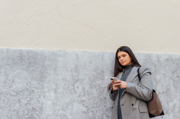 Hermosa mujer caucásica con su teléfono inteligente mientras se apoya en la pared