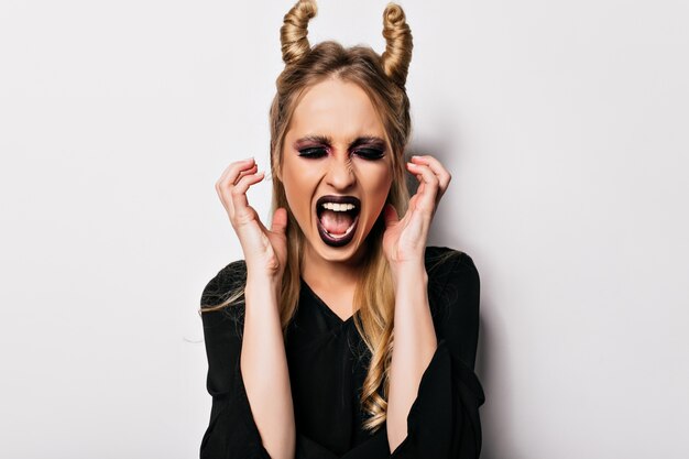 Hermosa mujer caucásica en traje de vampiro gritando en la pared blanca. Chica de moda en ropa negra jugando en la sesión de fotos de halloween.