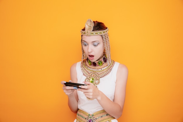 Hermosa mujer como cleopatra en traje egipcio antiguo jugando con smartphone sorprendido y sorprendido en naranja
