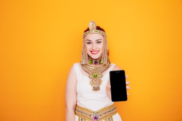 Hermosa mujer como Cleopatra en traje egipcio antiguo mostrando smartphone feliz y positivo sonriendo alegremente en naranja