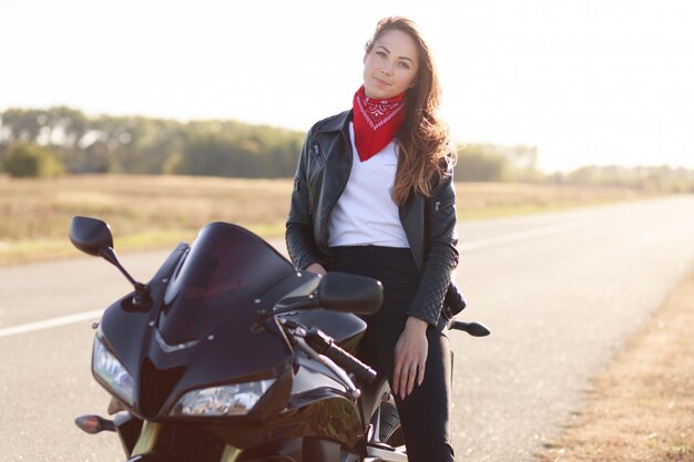 Hermosa mujer conductora se sienta en una moto negra rápida, una chaqueta de cuero vestida, viaja por el país en moto, se detiene al costado