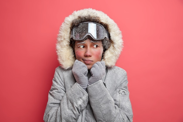 Hermosa mujer congelada tiembla de frío durante la nevada y la baja temperatura en febrero usa una chaqueta gris cálida y gafas de esquí para hacer snowboard en las montañas.