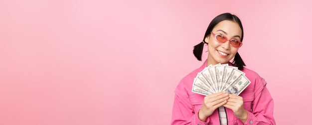 Hermosa mujer coreana con gafas de sol mostrando dólares dinero en efectivo sonriendo complacido concepto de préstamos rápidos microcrédito y pago de pie sobre fondo rosa