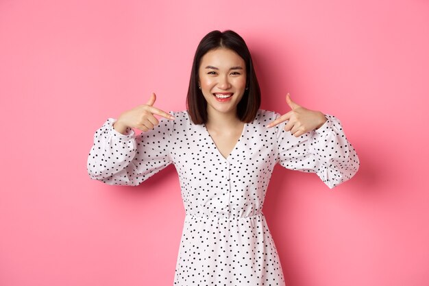 Hermosa mujer coreana señalando con el dedo a su logotipo y sonriendo, de pie en vestido sobre fondo rosa romántico