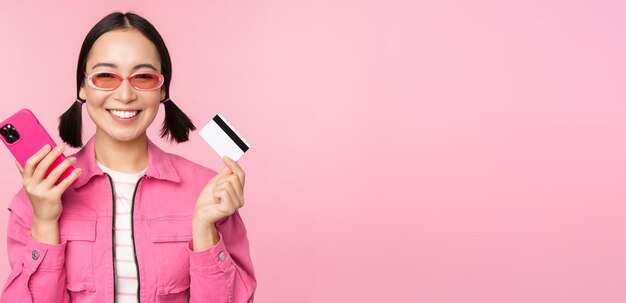 Hermosa mujer coreana sosteniendo una tarjeta de crédito de un teléfono inteligente sonriendo a la cámara comprando compras en línea con un teléfono móvil de pie sobre fondo rosa