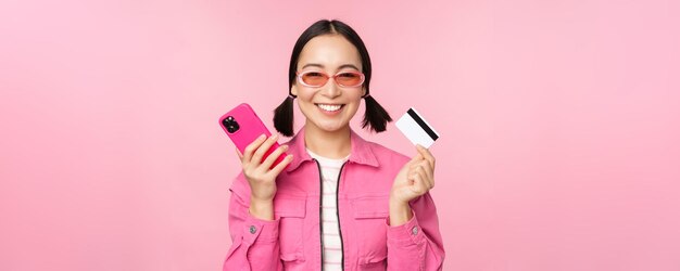 Hermosa mujer coreana sosteniendo una tarjeta de crédito de un teléfono inteligente sonriendo a la cámara comprando compras en línea con un teléfono móvil de pie sobre fondo rosa