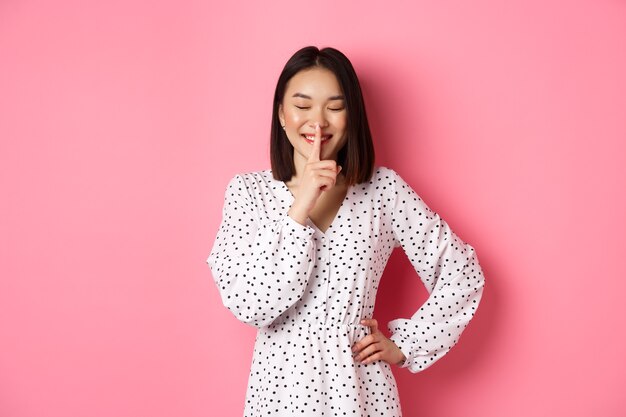 Hermosa mujer coreana en vestido de moda pidiendo mantener silencio en secreto con una sonrisa suave y los ojos cerrados ...