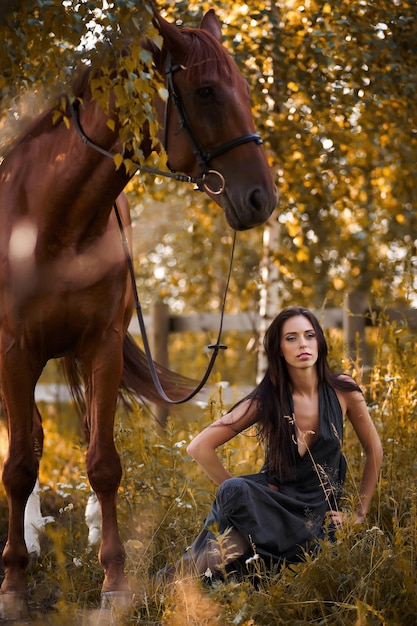 Hermosa mujer delgada sentada en el suelo con un caballo marrón cerca de ella.