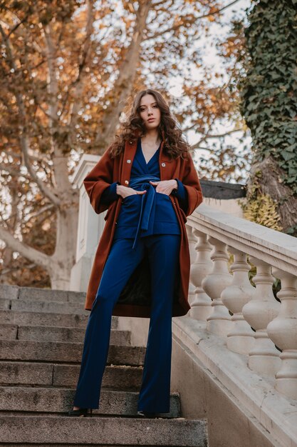 Hermosa mujer delgada sonriente con estilo con el pelo rizado caminando en las escaleras de la calle vestida con abrigo marrón cálido y traje azul, estilo callejero de moda de otoño