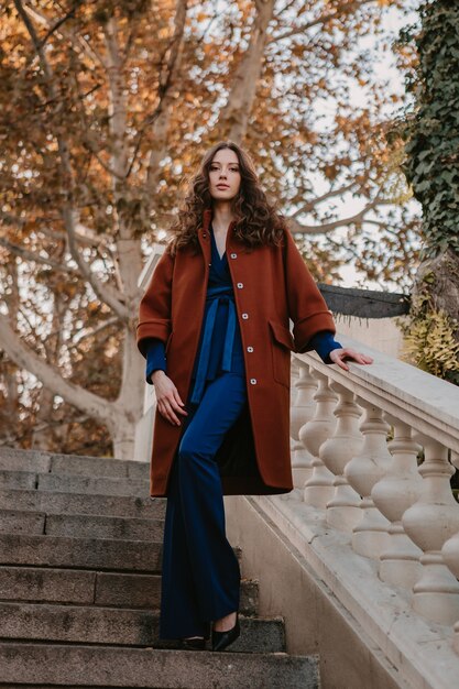 Hermosa mujer delgada sonriente con estilo con el pelo rizado caminando en las escaleras de la calle vestida con abrigo marrón cálido y traje azul, estilo callejero de moda de otoño