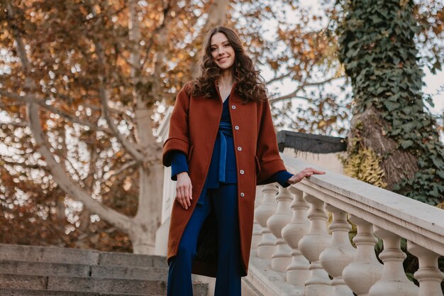 Hermosa mujer delgada sonriente con estilo con el pelo rizado caminando en las escaleras de la calle vestida con abrigo marrón cálido y traje azul, estilo callejero de moda de otoño