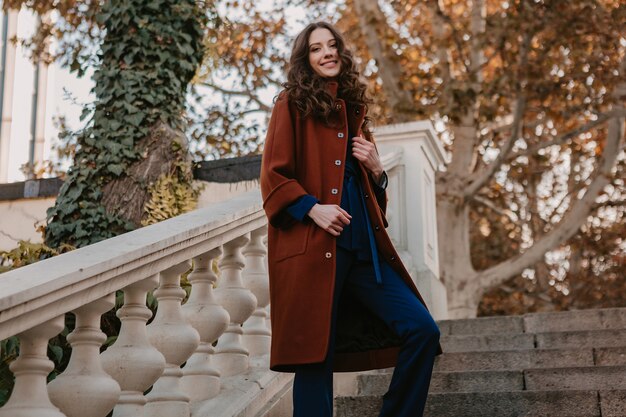 Hermosa mujer delgada sonriente con estilo con el pelo rizado caminando en las escaleras de la calle vestida con abrigo marrón cálido y traje azul, estilo callejero de moda de otoño