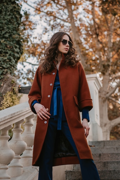 Hermosa mujer delgada sonriente con estilo con el pelo rizado caminando en las escaleras de la calle vestida con abrigo marrón cálido y traje azul, estilo callejero de moda de otoño