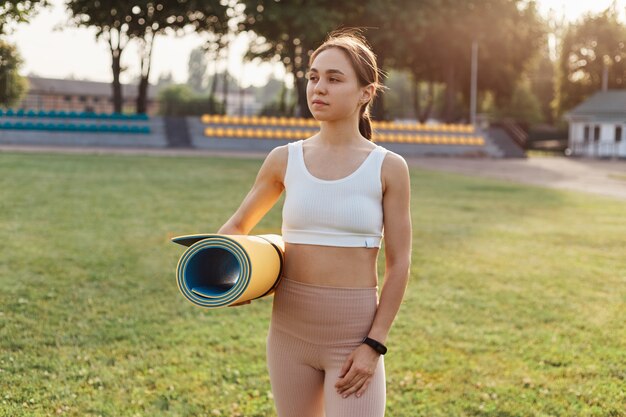 Hermosa mujer delgada vestida con leggins beige y top blanco posando al aire libre y mirando a otro lado, sosteniendo karemat en las manos, de pie en el estadio, tiene el cabello oscuro, haciendo ejercicio.