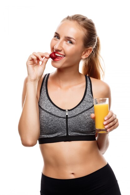 Hermosa mujer deportiva posando, sosteniendo un vaso de jugo y comiendo fresa