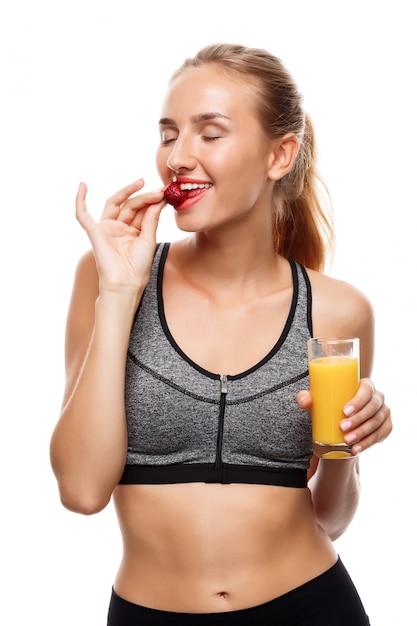 Hermosa mujer deportiva posando, sosteniendo un vaso de jugo y comiendo fresa