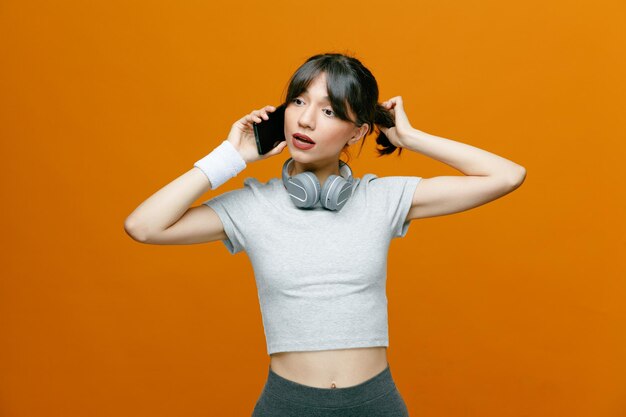 Hermosa mujer deportiva en ropa deportiva con auriculares hablando por teléfono móvil con aspecto seguro e intrigado de pie sobre fondo naranja