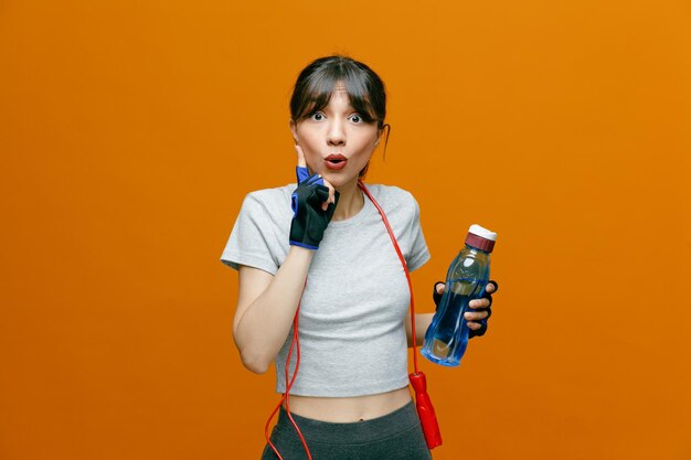 Hermosa mujer deportiva en ropa deportiva con saltar la cuerda en guantes sosteniendo una botella de agua mirando a la cámara feliz y sorprendida mostrando el dedo índice de pie sobre fondo naranja