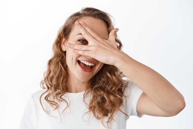 Hermosa mujer despreocupada mira a través de los dedos cubriendo los ojos con la mano y sonriendo feliz de pie en camiseta contra el fondo blanco.