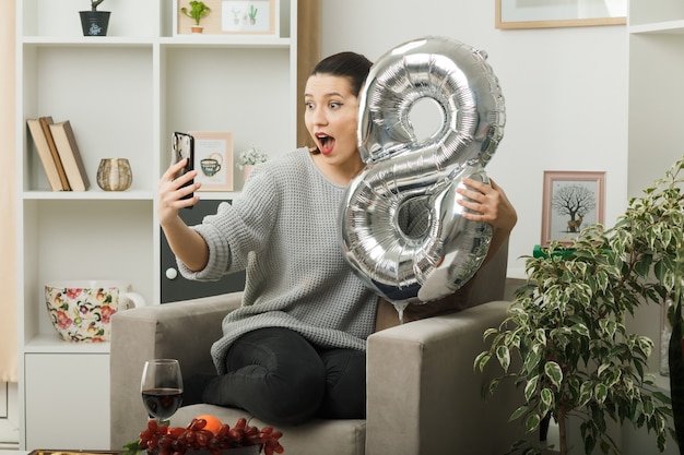 Hermosa mujer en el día de la mujer feliz sosteniendo el globo número ocho sentado en un sillón en la sala de estar