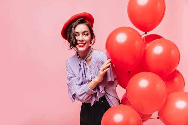 Hermosa mujer de elegante blusa y boina mira a la cámara con una sonrisa. Retrato de niña con labios rojos posando con globos.