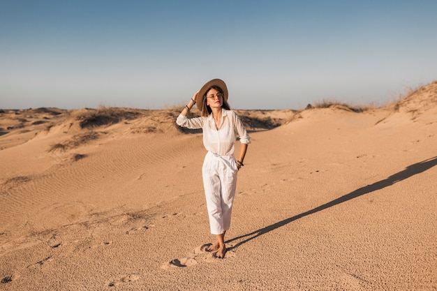 Hermosa mujer elegante caminando en la arena del desierto en traje blanco con sombrero de paja en la puesta del sol