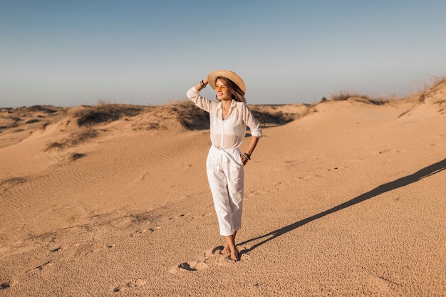 Hermosa mujer elegante caminando en la arena del desierto en traje blanco con sombrero de paja en la puesta del sol