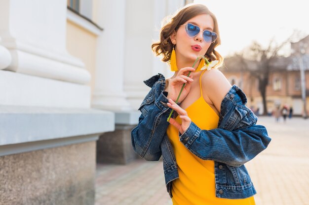 Hermosa mujer elegante hipster divirtiéndose, moda callejera, sosteniendo flores, vestido amarillo, chaqueta vaquera, estilo boho, tendencia de moda primavera verano, gafas de sol, sonriendo, soleado, coqueto