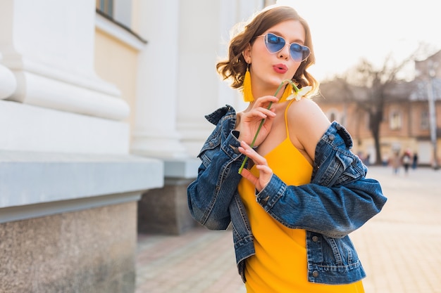 Hermosa mujer elegante hipster divirtiéndose, moda callejera, sosteniendo flores, vestido amarillo, chaqueta vaquera, estilo boho, tendencia de moda primavera verano, gafas de sol, sonriendo, soleado, coqueto