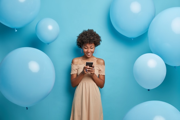 Hermosa mujer elegante mira felizmente el dispositivo de teléfono inteligente, sonríe agradablemente y lee buenas noticias en Internet, vestida con un vestido largo de verano, posa contra la pared azul decorada con globos