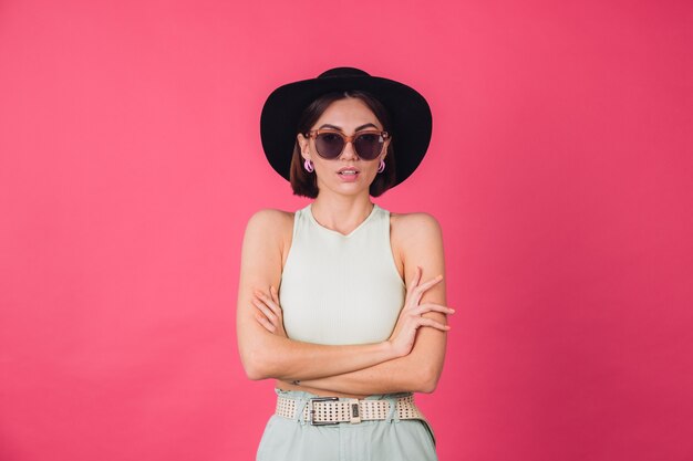 Hermosa mujer elegante con sombrero y gafas de sol posando sobre pared roja rosa