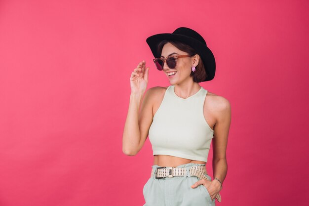 Hermosa mujer elegante con sombrero y gafas de sol posando sobre pared roja rosa