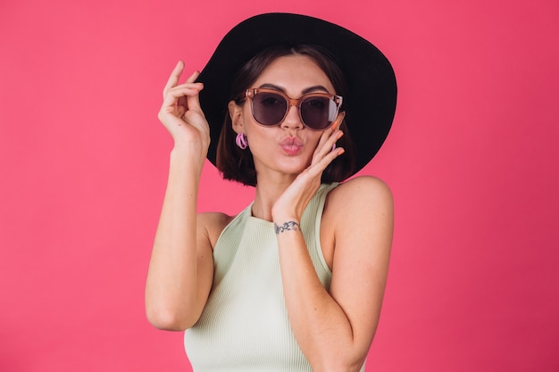 Hermosa mujer elegante con sombrero y gafas de sol posando sobre pared roja rosa