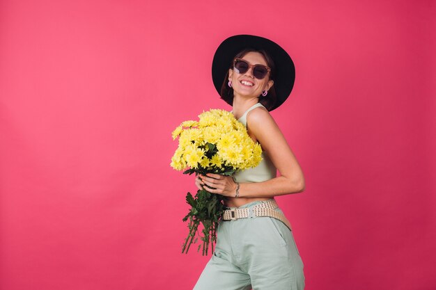 Hermosa mujer elegante con sombrero y gafas de sol posando, sosteniendo un gran ramo de ásteres amarillos, estado de ánimo primaveral, emociones positivas espacio aislado