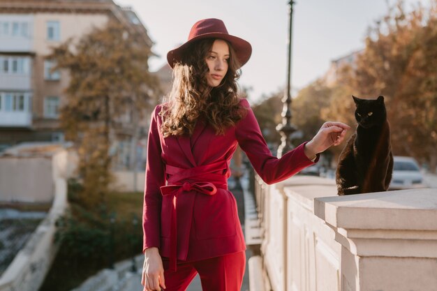 Hermosa mujer elegante en traje morado en las calles de la ciudad, tendencia de moda primavera verano otoño temporada con sombrero, mirando un gato