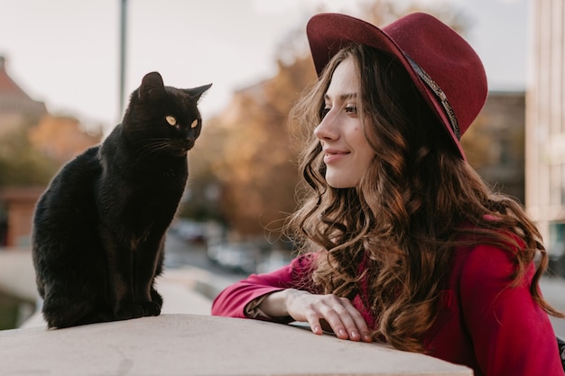 Foto gratuita hermosa mujer elegante en traje morado y sombrero caminando en las calles de la ciudad, tendencia de moda primavera verano otoño temporada, gato negro