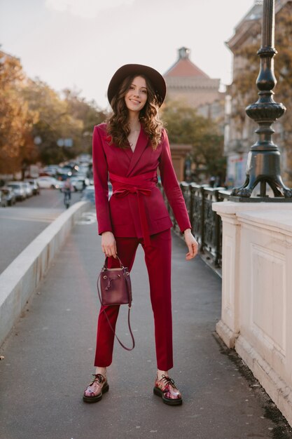 Hermosa mujer elegante en traje púrpura caminando en las calles de la ciudad, tendencia de moda primavera verano otoño temporada con sombrero, sosteniendo el bolso