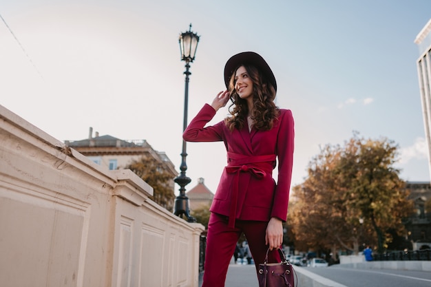 Hermosa mujer elegante en traje púrpura caminando en las calles de la ciudad, tendencia de moda primavera verano otoño temporada con sombrero, sosteniendo el bolso