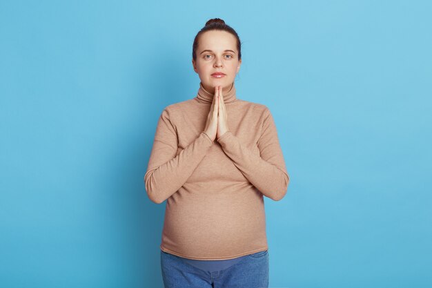 Hermosa mujer embarazada europea con las palmas juntas orando por su futuro bebé con ropa casual, se encuentra aislada en la pared azul, intenta relajarse antes de ir a la casa de maternidad.